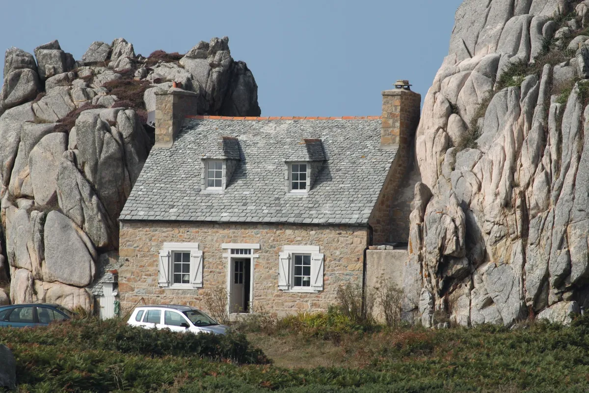 Longère en granit breton nichée entre de grands rochers de granit, toiture ardoise, voiture garée au premier plan, végétation dense autour