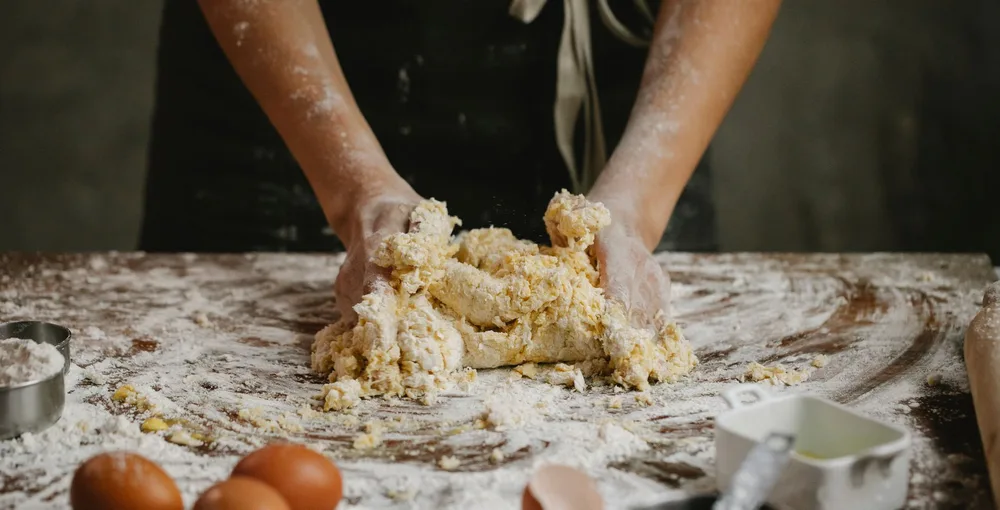 Mains pétrissant une pâte briochée sur un plan de travail fariné, oeufs visibles au premier plan, technique de pétrissage pour kouign-amann
